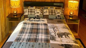A cozy bedroom with wooden walls, a bed with a 'Adventure' themed duvet, two bedside lamps, and three framed sunset pictures on the wall.