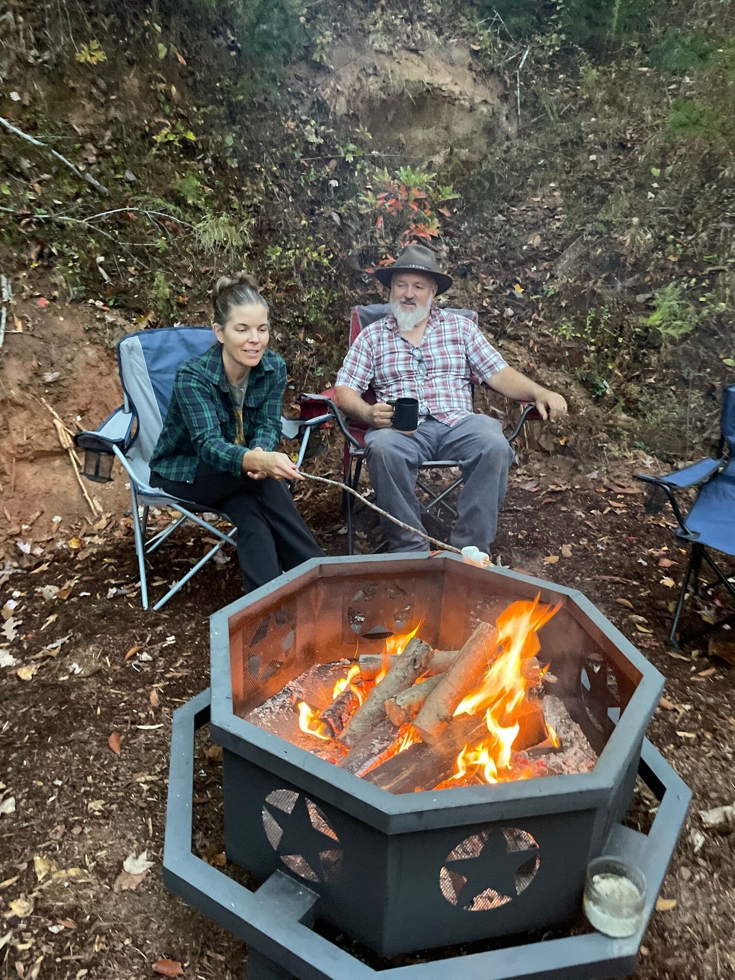 Dos personas sentadas en sillas alrededor de un pozo de fuego hexagonal con leña ardiendo.