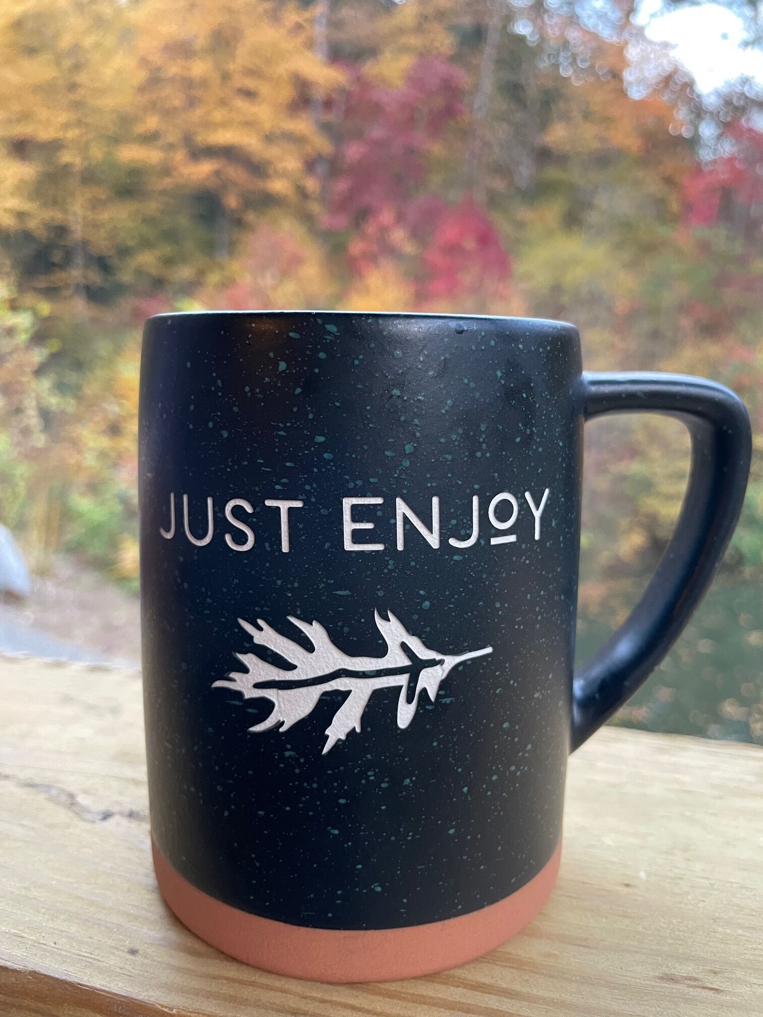 A black mug with "JUST ENJOY" and a leaf design on a wooden surface.
