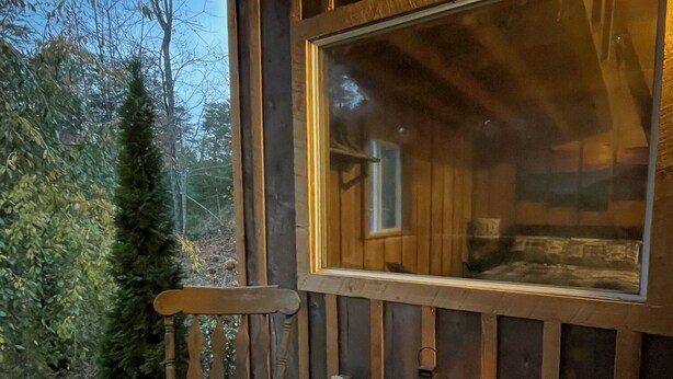 A wooden deck with a rocking chair and a bench, a lantern, and a window overlooking a forested area.