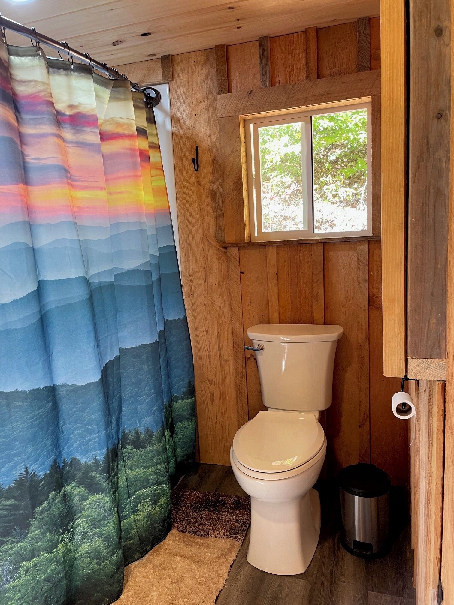 Une salle de bain avec un intérieur en bois, des toilettes, une fenêtre et un rideau de douche orné d’un motif représentant un paysage naturel.