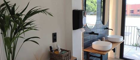 A modern bathroom with a large mirror, two round sinks, a potted plant, a small table with a basket, and a trash bin.