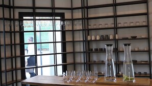 Une pièce comprenant une table en bois sur laquelle sont exposés divers objets en verre, une étagère en métal noir où sont rangés des verres et des bocaux, et une fenêtre donnant sur une clôture verte et sur une personne se trouvant à l’extérieur.