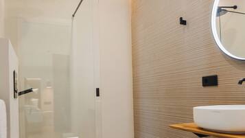 Une salle de bain moderne avec une douche à parois de verre, un plancher en bois, des toilettes blanches, un lavabo avec un comptoir en bois et un miroir rond.