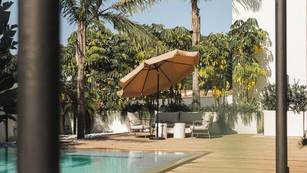 Un espace piscine agrémenté de chaises longues et de parasols, entouré de palmiers et d’un bâtiment blanc.
