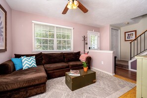 A living room with a brown sofa, a window with blinds, a ceiling fan, and a staircase. - Spacious 3-Bedroom, 1.5-Bath Home in Central Maryland with Screened Porch & More (Severn)