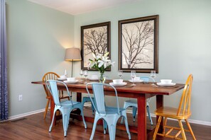 A dining area with a wooden table set for four, surrounded by four chairs, two framed artworks on the wall, and a vase with flowers on the table. - Spacious 3-Bedroom, 1.5-Bath Home in Central Maryland with Screened Porch & More (Severn)
