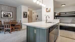Una cocina compacta con una isla central, un comedor con sillas de madera, y una cama individual en la habitación contigua.