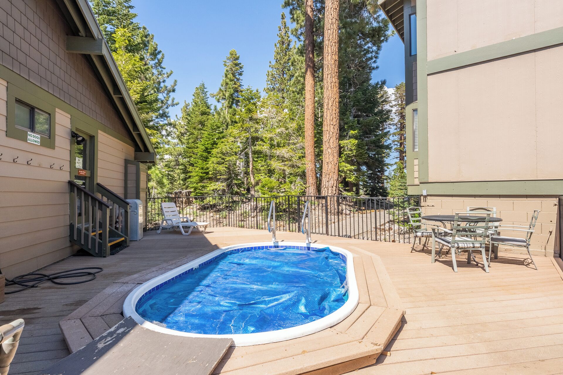A swimming pool with a blue cover, surrounded by a wooden deck and a metal fence. There are lounge chairs and a table with chairs on the deck.