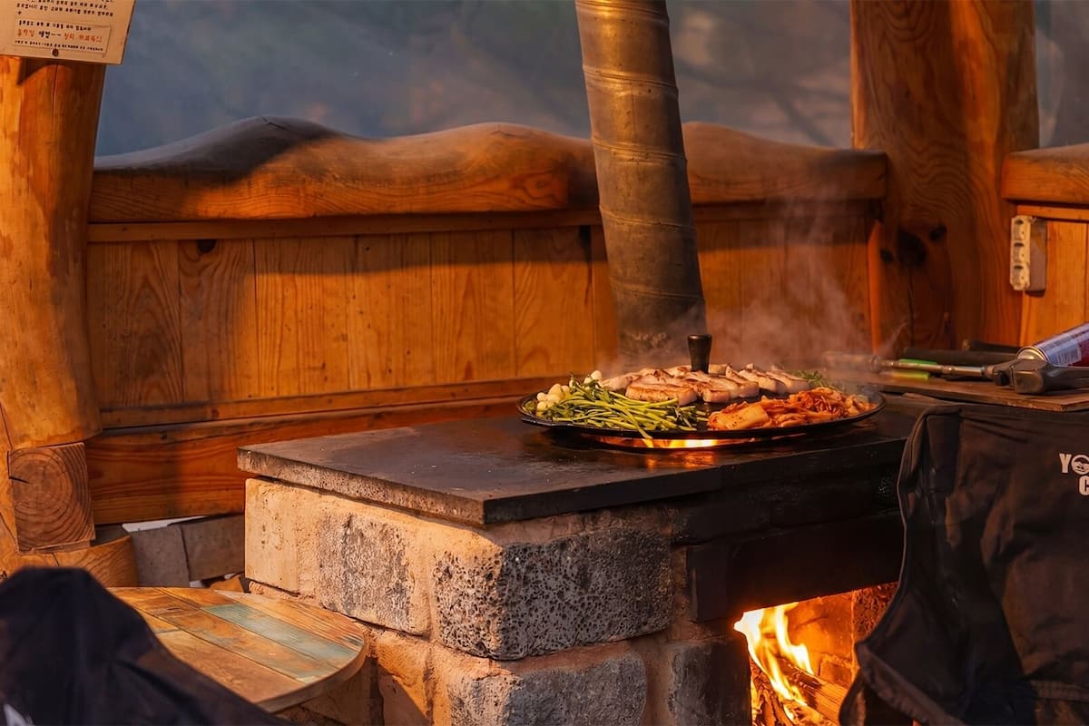 A wood-fired grill with food, a stone stove, and a 'YOUNKER CAMP' bag.