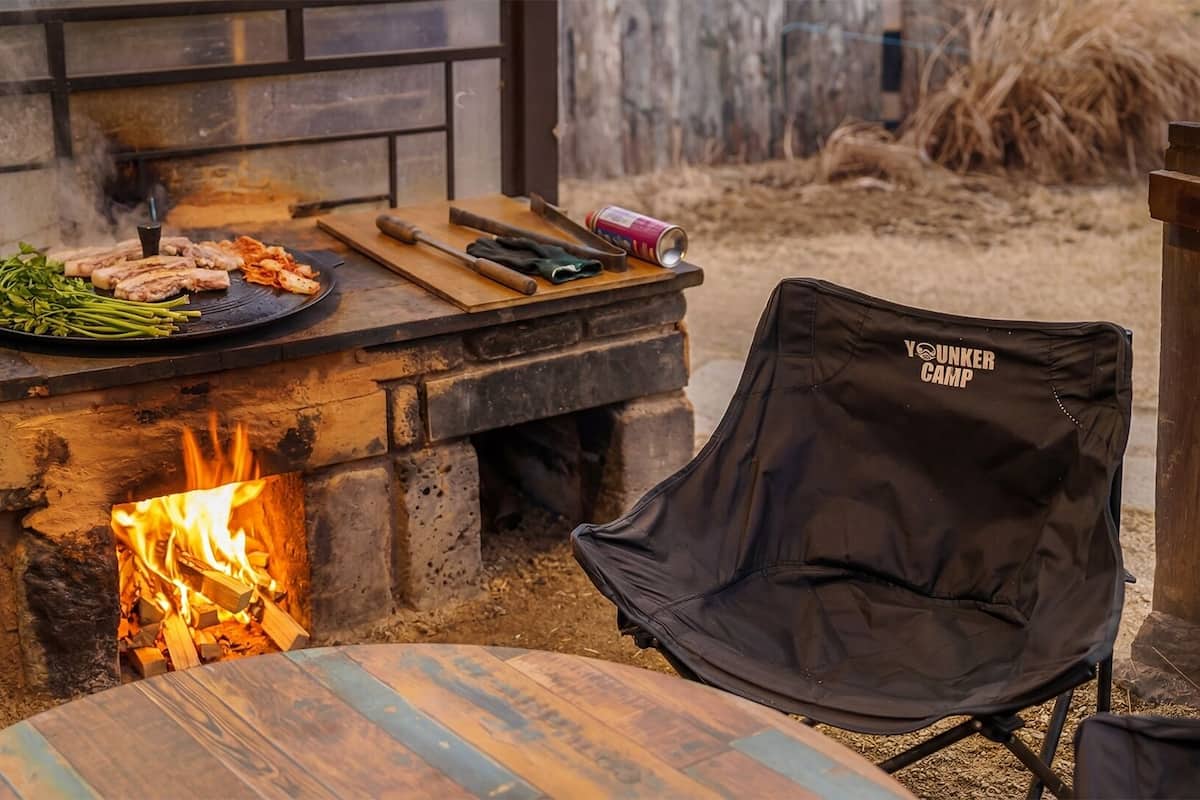 A stone fire pit with a grill above it, a chair, and a table with a firewood log.