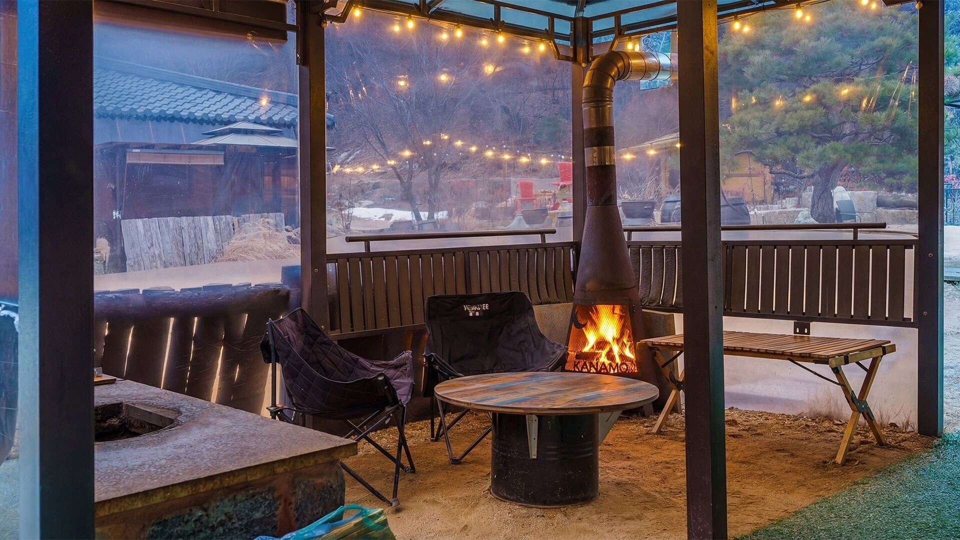 Ein Sitzbereich im Freien mit einem Holzofen, Stühlen, einem Tisch und Blick auf ein traditionelles Gebäude.