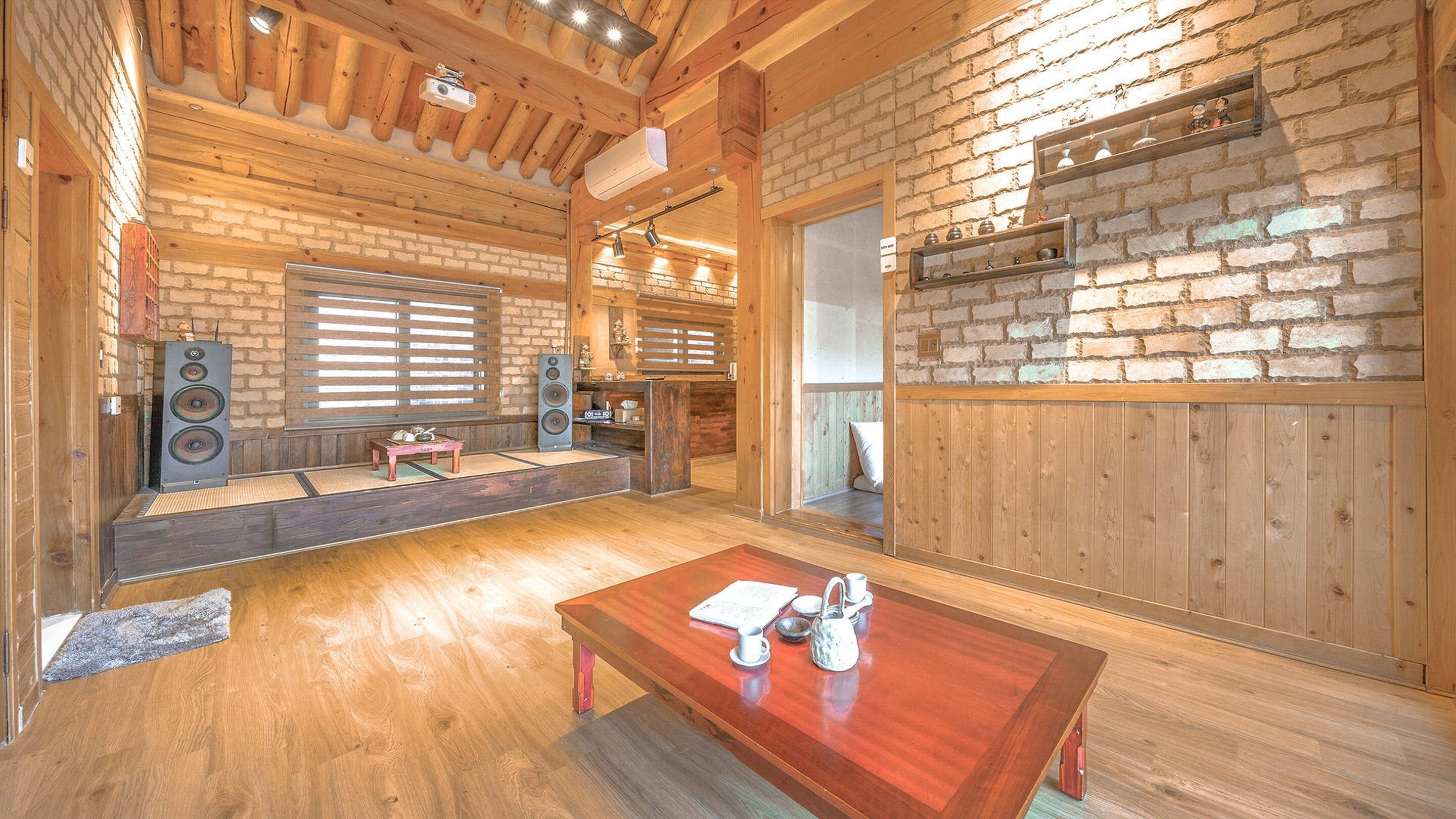 A spacious room with a wooden ceiling, brick walls, and a large window. There is a wooden table with a teapot and cups, and a seating area with a bench and cushions.