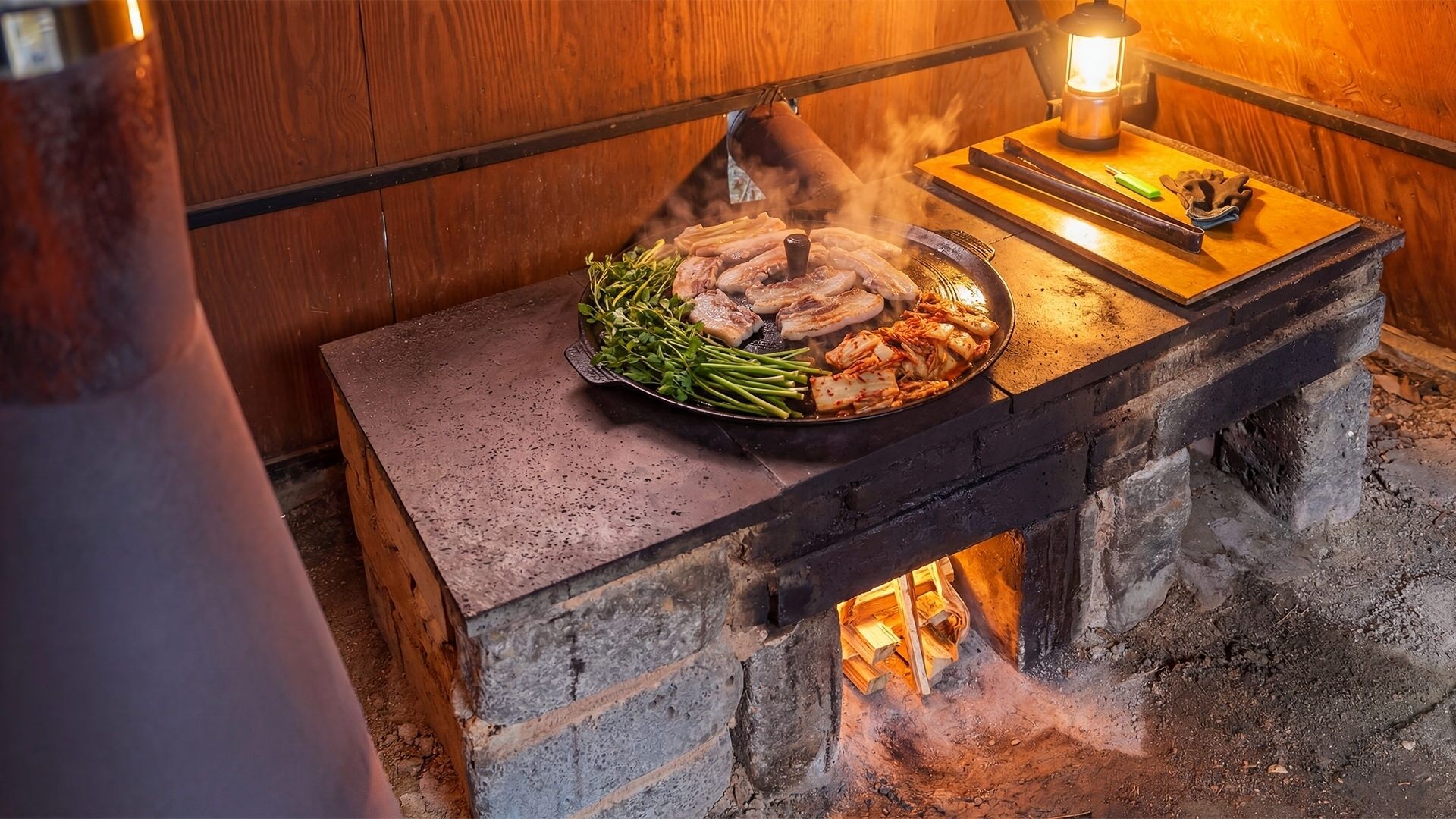Un barbecue coréen traditionnel, avec un gril, des légumes et de la viande servis sur une table en pierre, une lanterne et une bouteille de sauce à proximité.