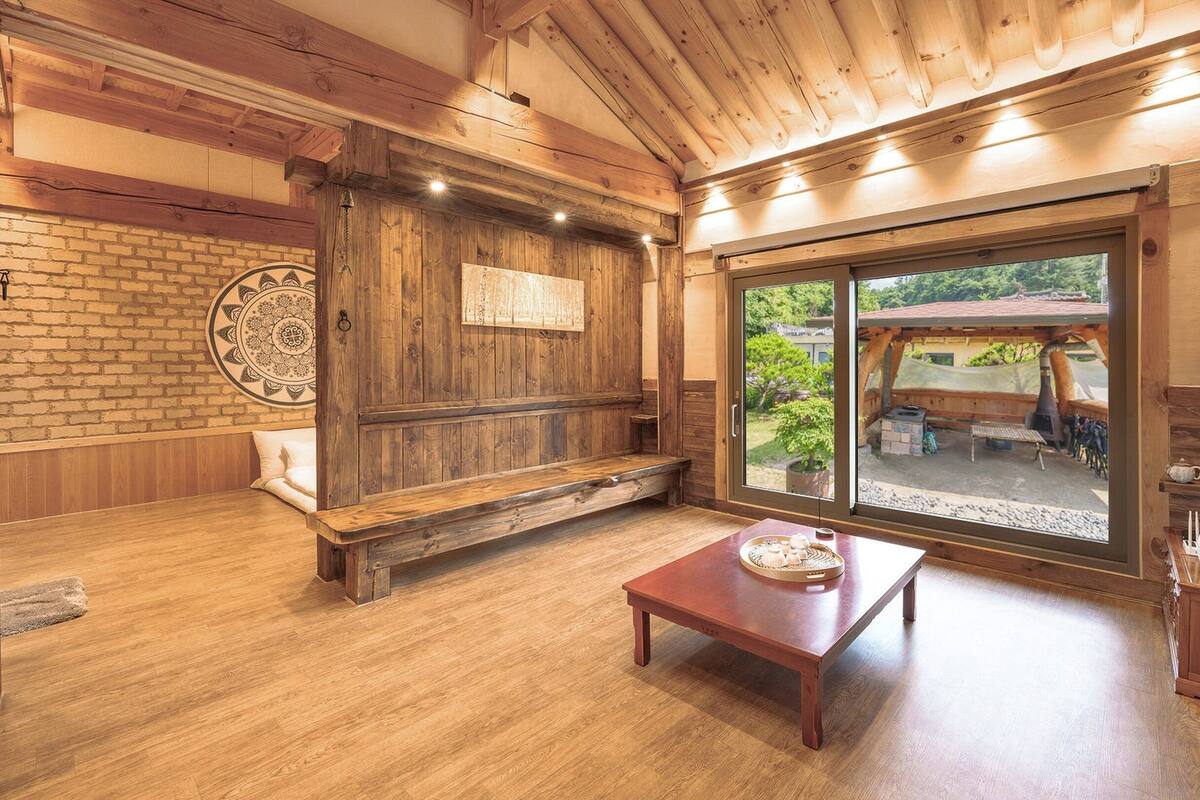 A rustic wooden interior with a bench, a table, and a large window overlooking a garden.