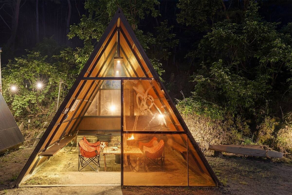 A triangular glass structure with a table and chairs inside, surrounded by trees.