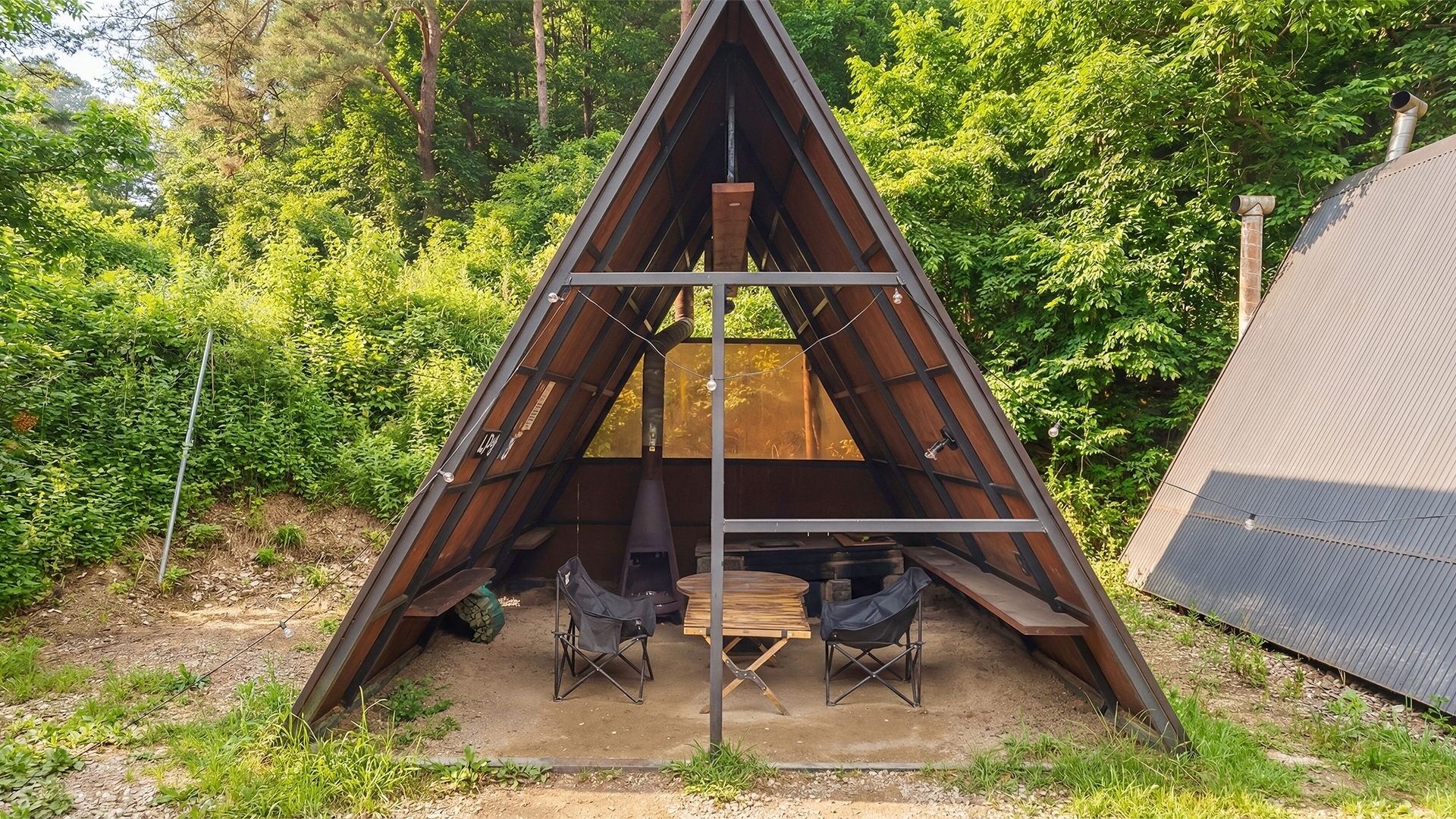 Eine dreieckige Metallkonstruktion mit Tisch und Stühlen im Inneren, umgeben von Grünpflanzen.