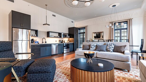 Un salon moderne comprenant un canapé, un pouf et une table basse, une cuisine avec des meubles sombres et des appareils électroménagers en acier inoxydable, et un coin repas avec une table et des chaises.