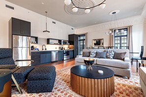 A modern living room with a sofa, ottoman, and coffee table, a kitchen with dark cabinetry and stainless steel appliances, and a dining area with a table and chairs. - 3BR Historic Dance Hall & Canal St Views | The Mayfair (New Orleans)