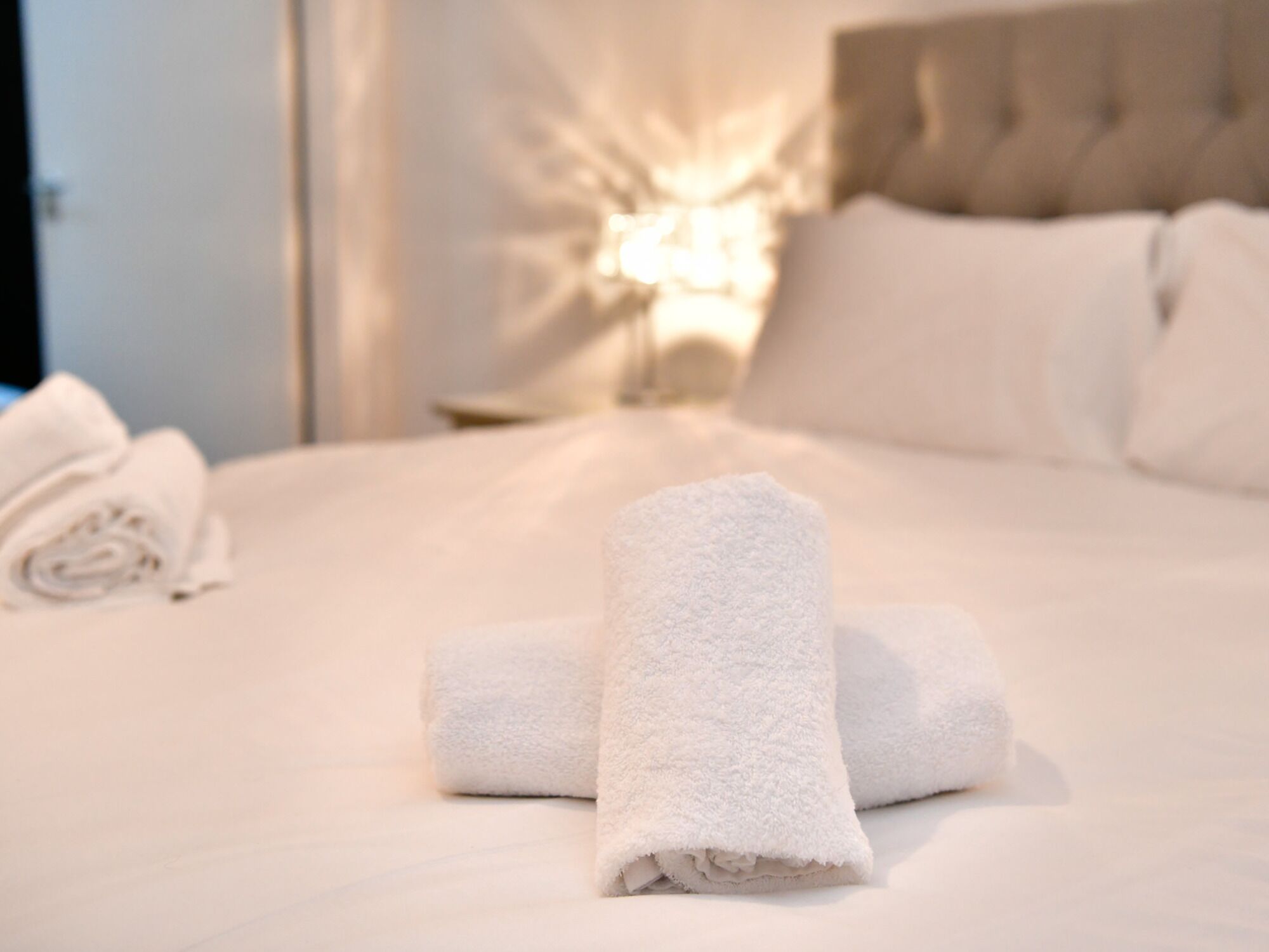 A neatly made bed with white linens and rolled towels, a headboard, and a bedside lamp.