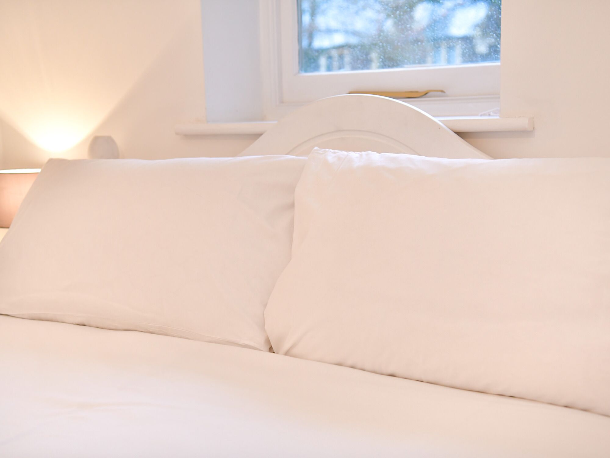 Un lit avec une literie blanche et une tête de lit, placé devant une fenêtre donnant sur une maison.
