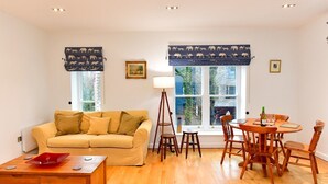 A living room with a beige sofa, wooden coffee table, and a dining area with a wooden table and chairs. There are two windows with blue curtains featuring animal prints. - Bright & Perfectly Located! - Pass the Keys (Cardiff)