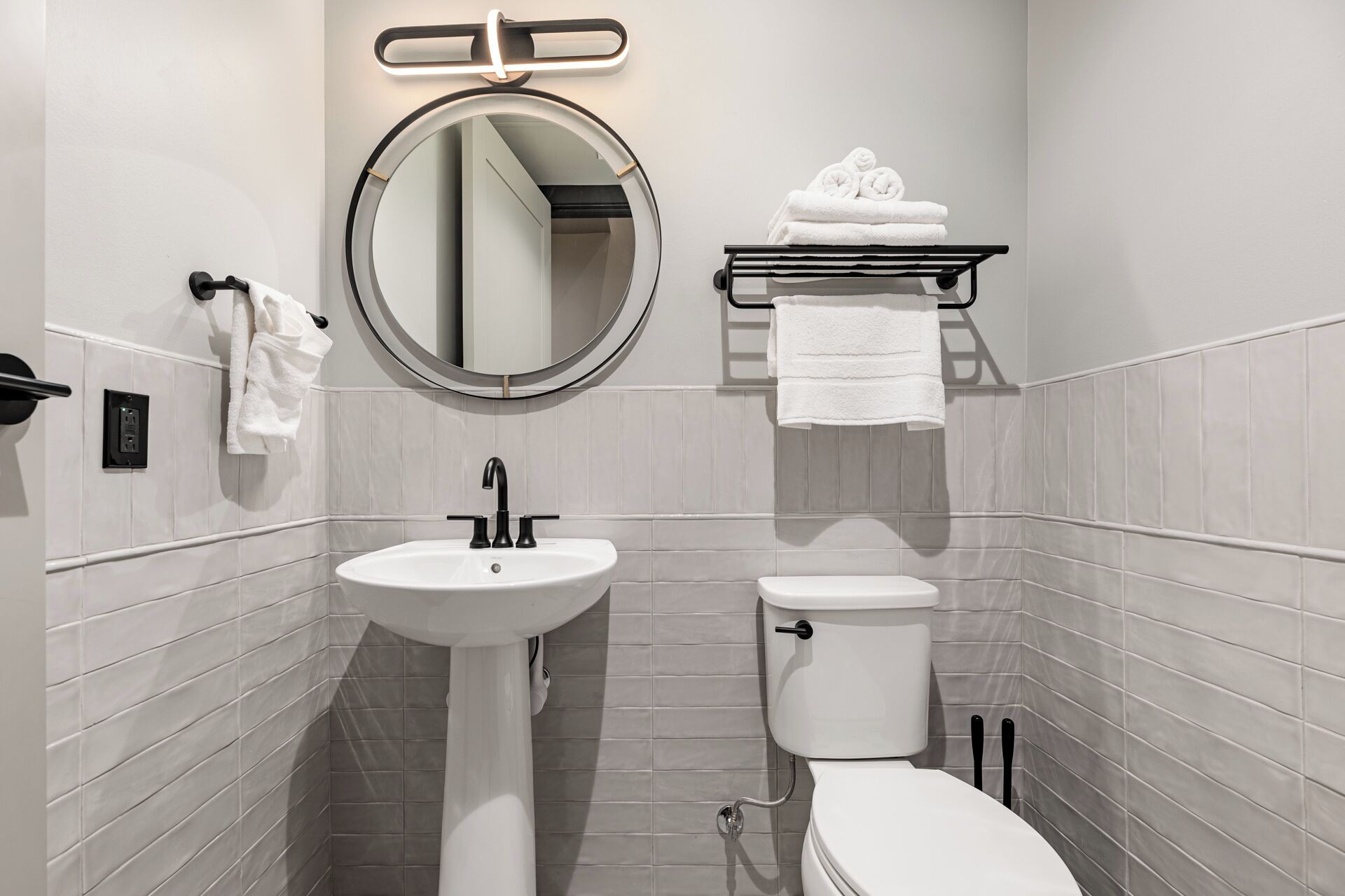 A modern bathroom with a pedestal sink, a toilet, a round mirror, and a towel rack with folded towels.