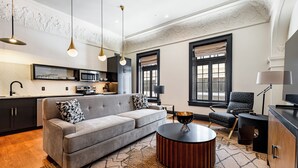 A modern living room with a grey sofa, a round coffee table, and a black armchair. There is a kitchen area with a microwave and a sink.