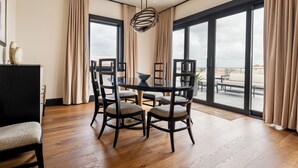 Un comedor moderno con una mesa redonda, cuatro sillas y una lámpara decorativa. Amplias ventanas con cortinas ofrecen vistas del paisaje urbano.
