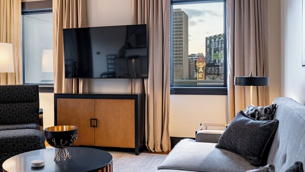 A modern hotel room with a flat-screen TV, a sofa, a coffee table, and a view of a cityscape through large windows. - 4BR Exclusive Downtown Group Penthouse | The Mayfair (New Orleans)