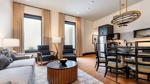 Ein modernes Wohnzimmer mit einer Couch, Sesseln, einem Couchtisch und einem Essbereich, im Hintergrund ist eine Küche zu sehen.