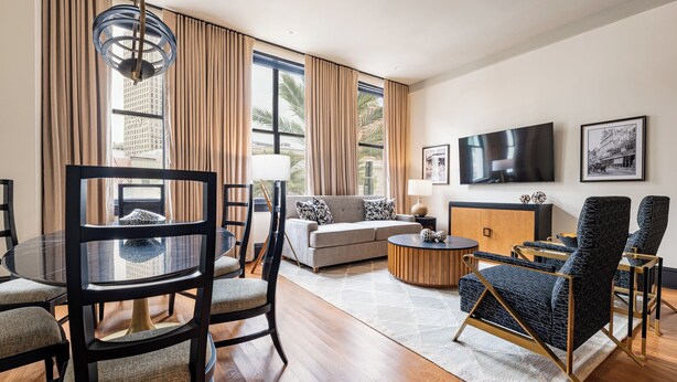 A modern living room with a sofa, a dining table with chairs, a TV mounted on the wall, and a large window with curtains. - 4BR Group Getaway Walk to French Quarter | The Mayfair (New Orleans)