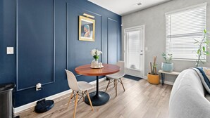 A modern living room with a dark blue accent wall, a round wooden table with two white chairs, a small side table with a vase and plants, and a sofa with a blue throw. - Electric & Vibrant home 15 min from Downtown ATL (Decatur)