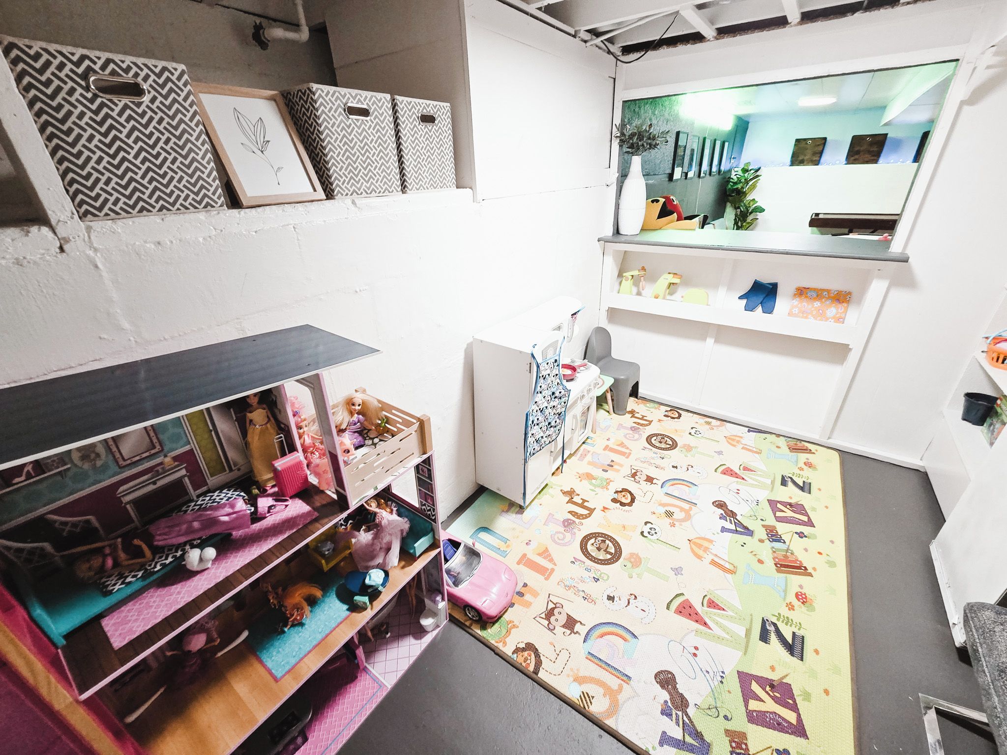 Une petite pièce comprenant une maison de poupée, un tapis à motifs et une fenêtre donnant sur des immeubles.