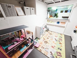Quarto compacto com uma casa de bonecas, um tapete estampado e uma janela com vista para edifícios.