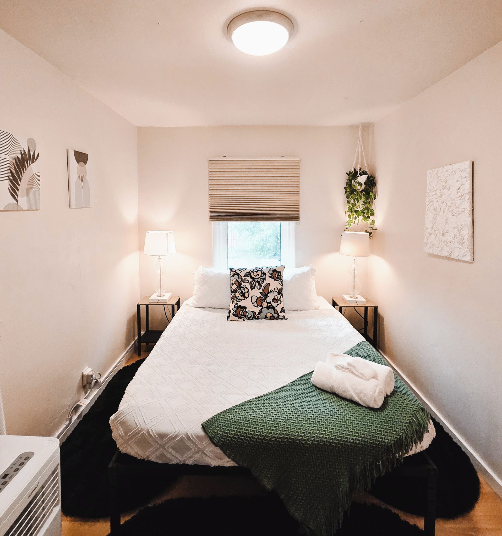 Een hotelkamer met een bed, nachtlampjes, een raam met zonwering, een hangplant en kunstwerken aan de muur.