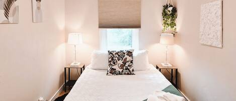 Habitación de hotel con cama, lámparas de noche, ventana con persianas, planta colgante y obras de arte en las paredes.