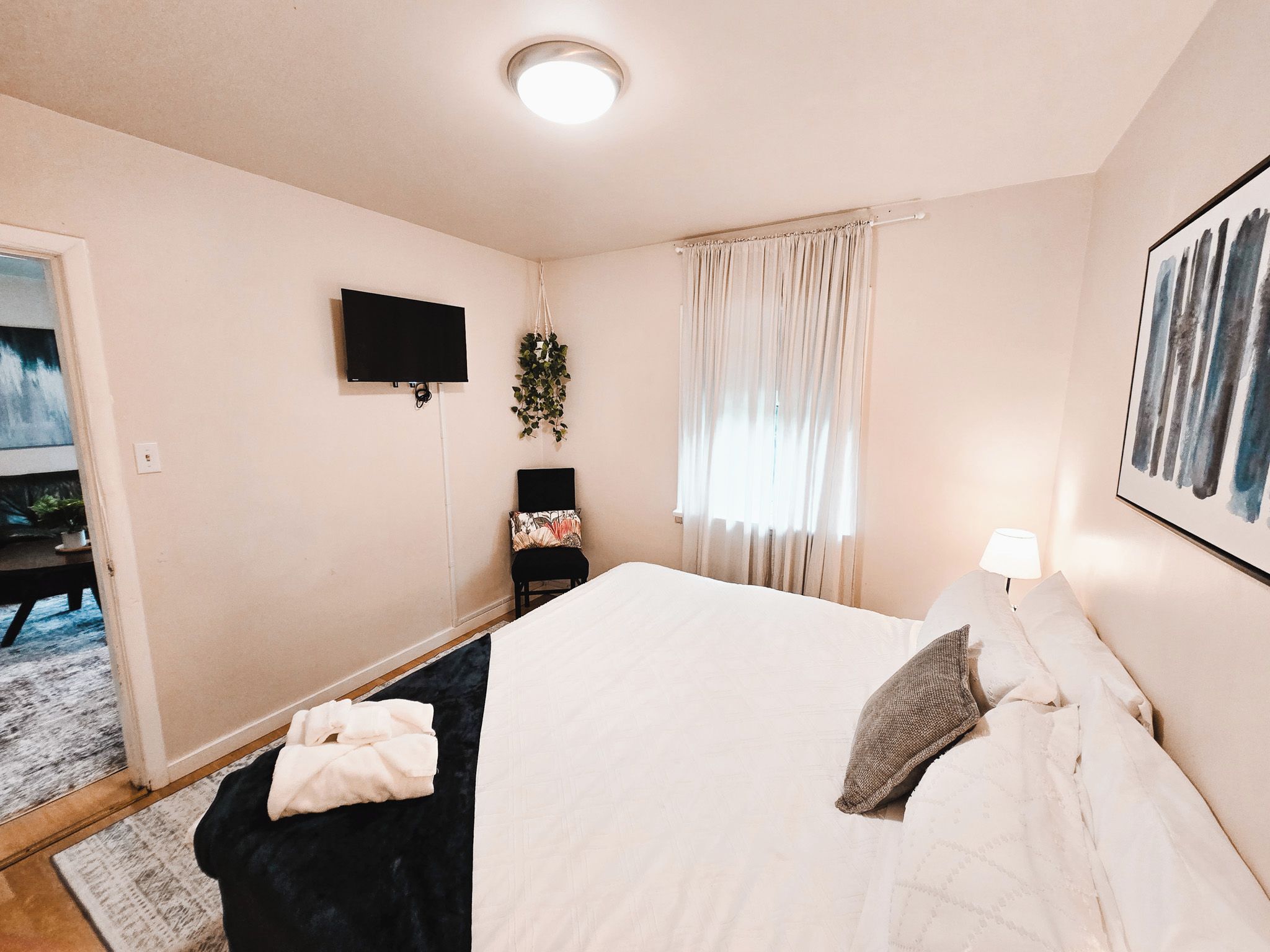 Een hotelkamer met een bed, een aan de muur bevestigde televisie, een raam met gordijnen, een nachtkastje met een lamp en een schilderij aan de muur.