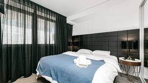 A modern hotel room with a large bed, a bedside table with a lamp, and a view of the outdoors through sheer curtains.