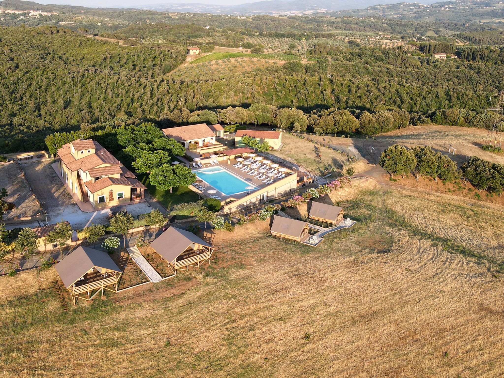 Vue aérienne d’un complexe hôtelier doté d’une piscine, de plusieurs bâtiments et entouré de collines boisées.