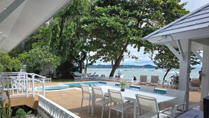 Un espace aménagé au bord de la piscine, avec des chaises blanches et un barbecue, donnant sur une étendue d’eau où se trouvent des bateaux.