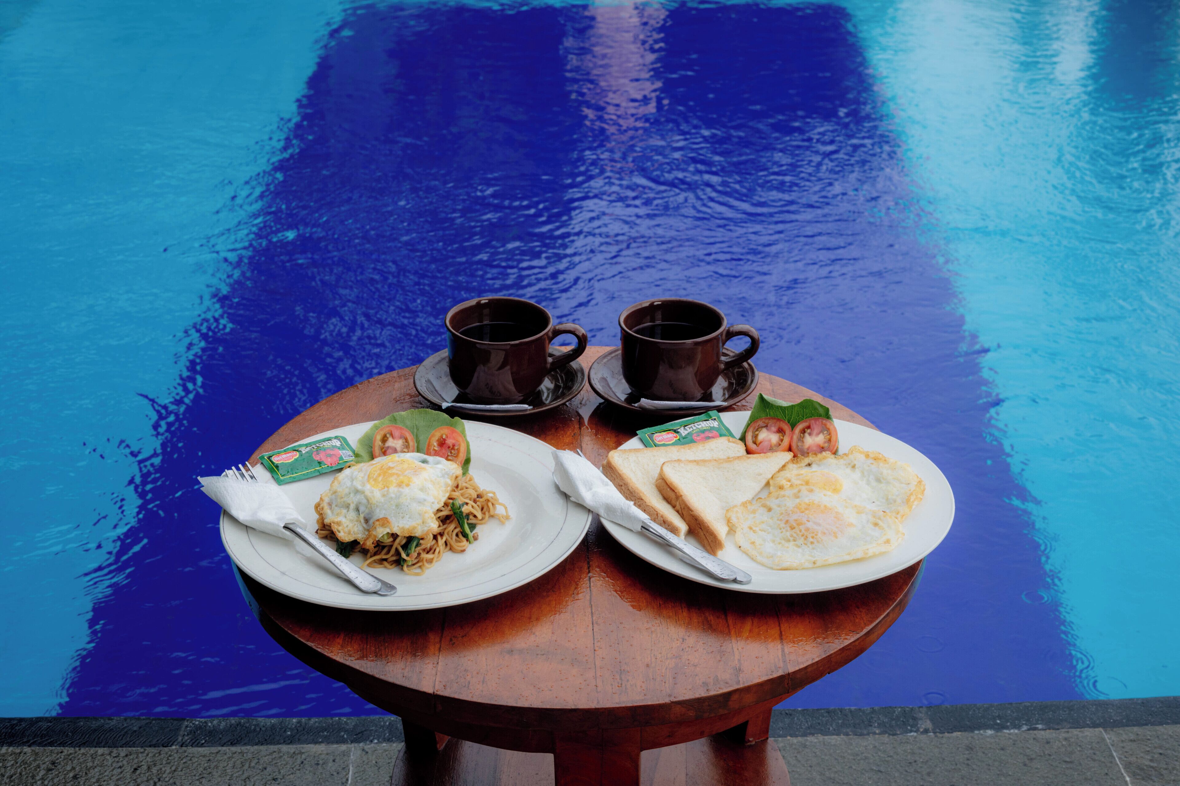 A wooden table with two plates of food and two cups of coffee by a pool.