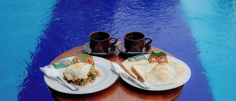 A wooden table with two plates of food and two cups of coffee by a pool.