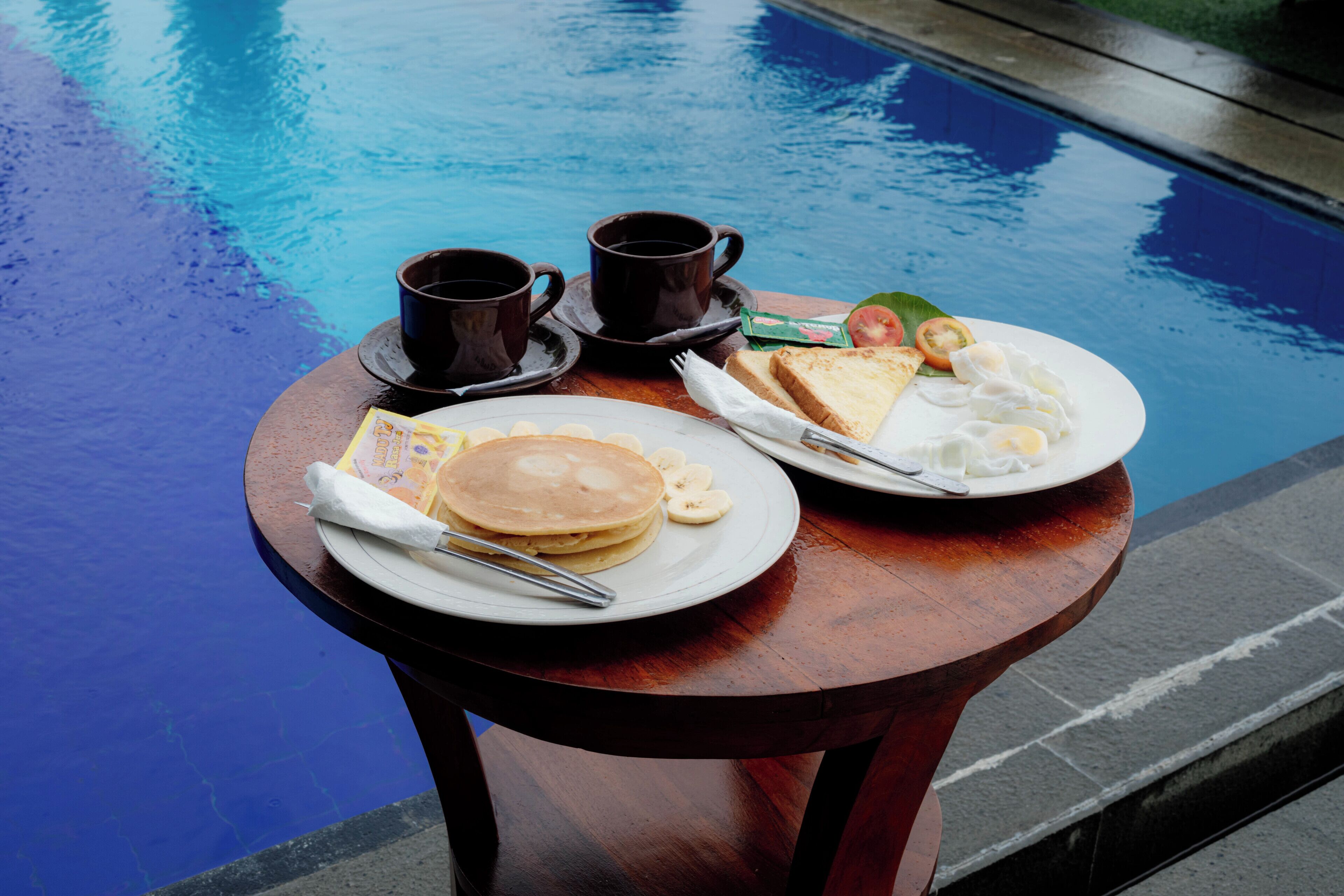 A breakfast table with pancakes, eggs, toast, and coffee by a pool.