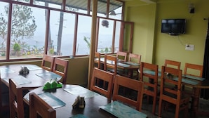 A dining area with wooden tables and chairs, large windows with a view of trees and mountains, and a mounted television on the wall. - Binsar View Homestay (Almora)