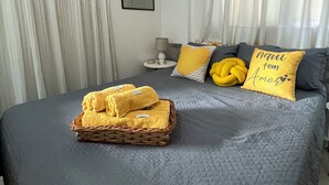 En pænt redt seng med et gråt tæppe, en kurv med foldede håndklæder og dekorative puder med hjerte-motiv.