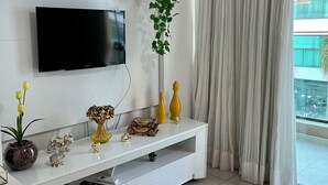 En moderne stue med et fladskærms-tv, et hvidt tv-møbel med dekorative genstande, en potteplante og udsigt til en bygning gennem lette gardiner.