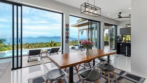 Een moderne keuken met uitzicht op de oceaan, een eethoek met een houten tafel en stoelen, en een grote schuifpui die leidt naar een balkon met een zwembad.