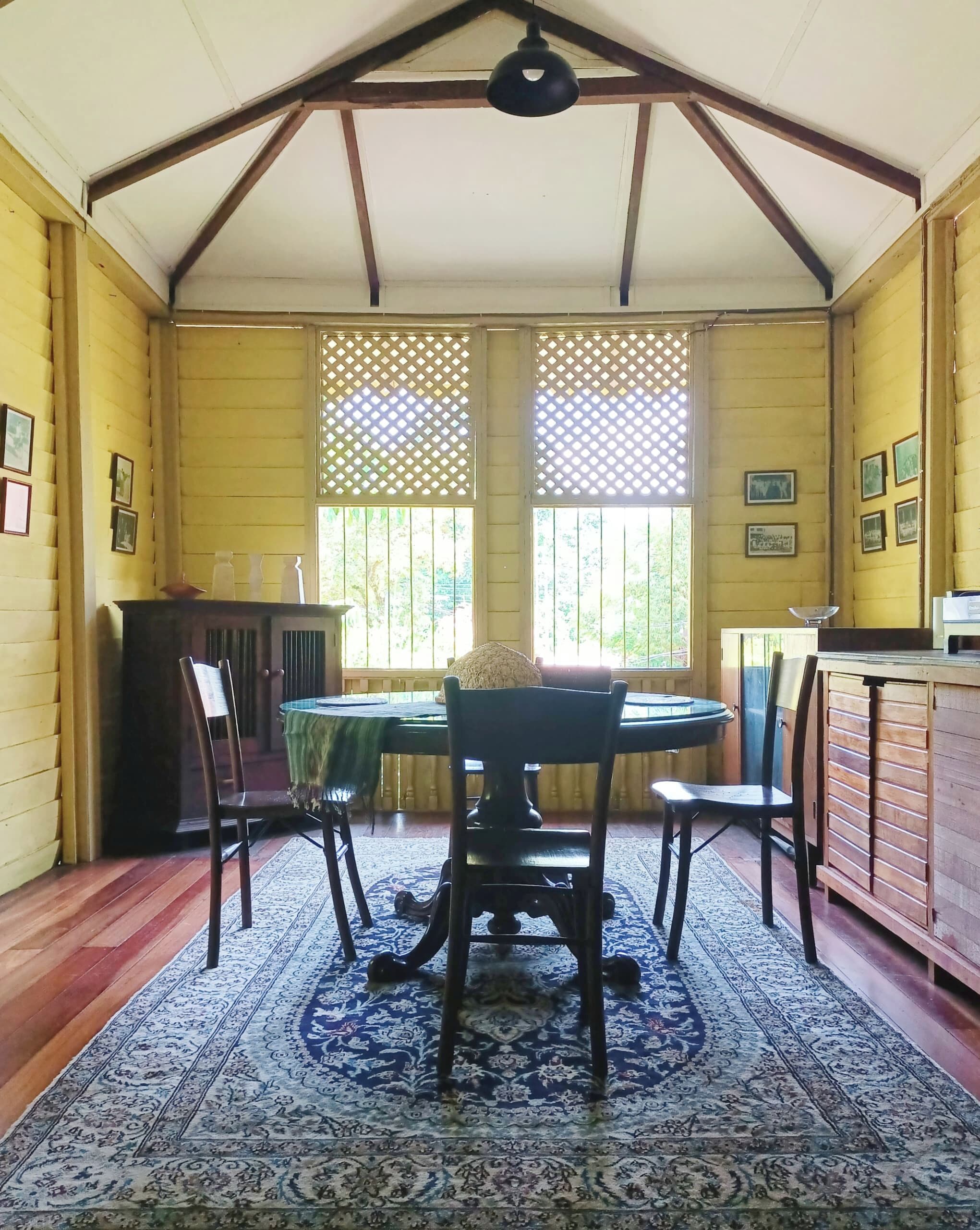 A dining area with a round table, chairs, a patterned rug, wooden walls, and windows with lattice screens.