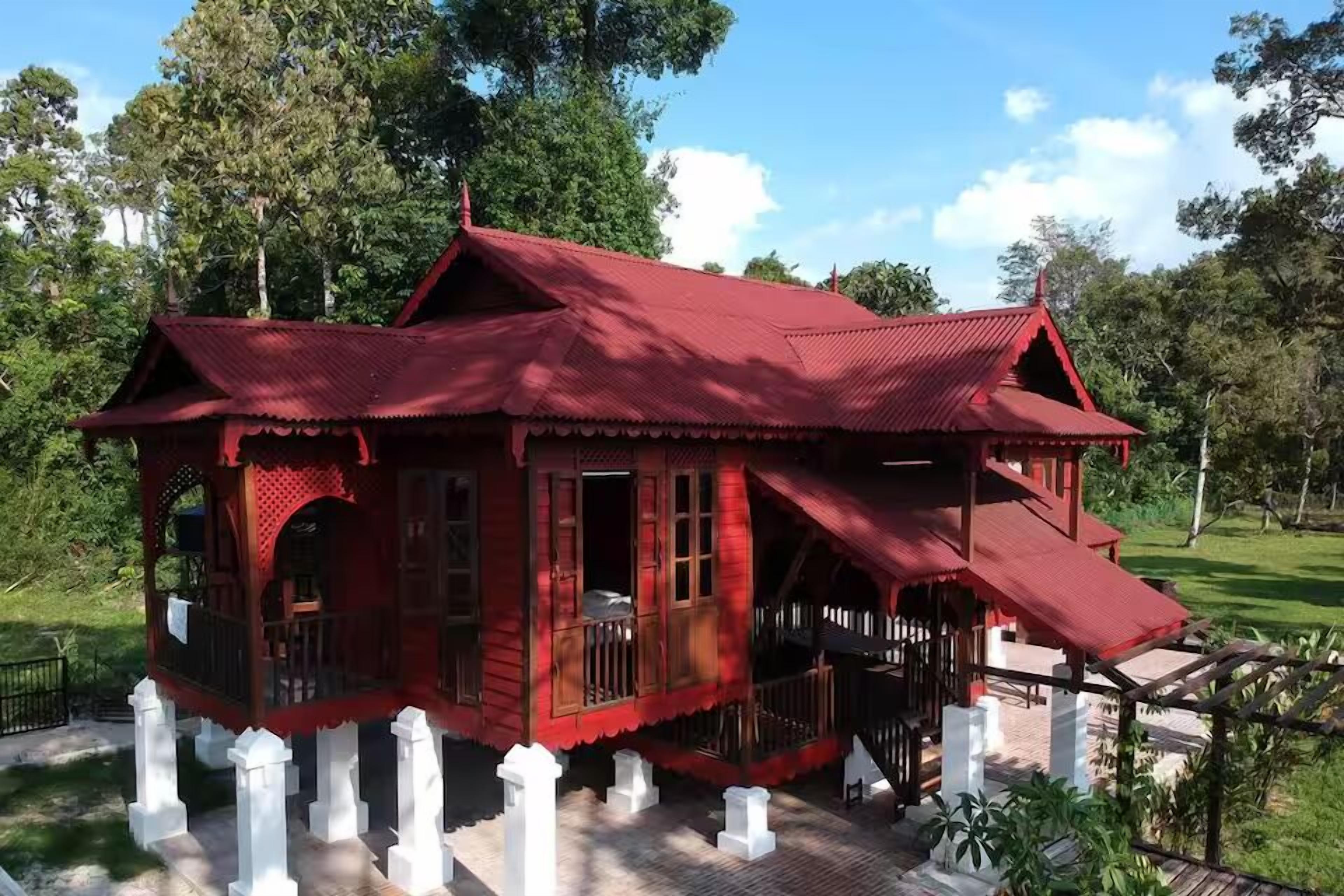 A traditional red wooden house with a balcony, surrounded by white pillars and lush greenery.
